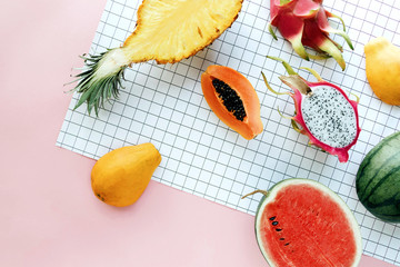 Flat lay tropical summer fruit on a white grid and pastel pink background. Creative food concept with pineapple, watermelon, Hawaii papaya, dragon fruit