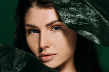 Wall Mural - attractive woman with freckles on face posing with palm leaves isolated on green