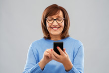Technology And Old People Concept - Smiling Senior Woman In Glasses Using Smartphone Over Grey Background