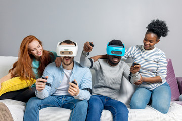 Two multiracial young couples having fun while visiting entertainment center in weekend. Emotional guys in VR headsets experience joy and excitement while driving virtual race car