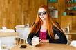 © Julio Ricco - Beautiful redhead model with dark glasses holding coffee cup in cafeteria. Cellphone on the table with blurred background.
