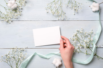 Womens hand holding blank paper card on light blue wooden background. Rustic table with flowers, wedding invitation card, letter, greeting card for Womens day. Trendy minimal design, Flat composition