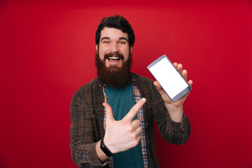 Wall Mural - Portrait of a confident young bearded man, showing blank screen mobile phone, pointing finger