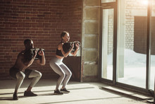 Young Mixed-race Fit Couple Doing Squats Exercise, While Holding In Hands Heavy Sandbag During Group Circuit Functional Training At The Well-lit Gym With Panoramic Windows.