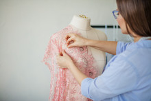 Skillful Fashion Designer Stands In A Dressmaker Studio Near The Mannequin And Fixes Lace Fabric On It Using Needles. Creation Or Modelling Of Fashion Clothes. Designer Clothes Concept