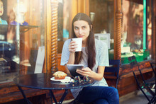 Young Female In Cafe Drinking Outdoors, Muffin On Table, Shop Window With Sun Reflection In Background 
