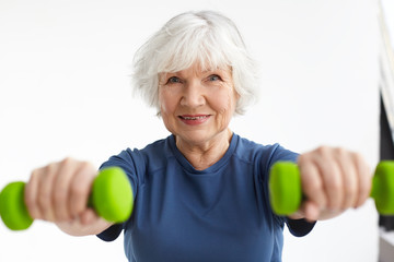 Active energetic happy elderly Caucasian female with gray hair enjoying physical exercises indoors, training at home using dumbbells, smiling broadly at camera. Selective focus on woman's face