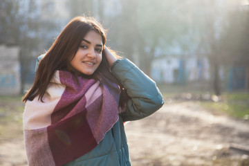 happy young fasion woman standing on the street in winter coat and big scarf touching her shiny brown hair  in the sunny day