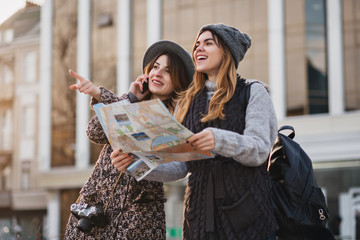 Happy travel together of two fashionable girls in sunny city centre. Young joyful women expressing positivity, using map, vacation with bags, camera, making photo, cheerful emotions, good day