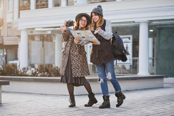 Stylish city portrait of two fashionable girls walking in Europe modern city centre. Fashionable friends travelling with backpack, map, camera, making photo, tourist, get a lost, place for text