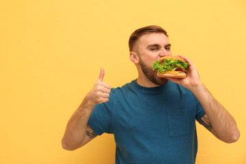 Wall Mural - Young man eating tasty burger on color background. Space for text