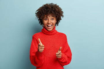 Wall Mural - Hey you. Cheerful dark skinned lady points at camera, says you are what we need, makes finger gun gesture, picks canditate, has broad smile, wears casual red outfit, isolated over blue background