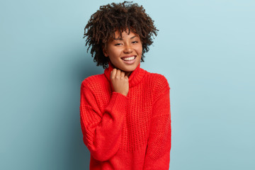 Wall Mural - Close up shot of gentle woman with satisfied expression, appealing appearance, keeps hand on collar of red sweater, smiles broadly, has white perfect teeth, isolated over blue background. Emotions