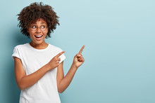 Beautiful Black Woman Looks To Side, Shows Free Advertising Space, Smiles Broadly, Dressed In White Casual T Shirt, Isolated Over Blue Background, Wears Spectacles For Good Vision. Its For Your Ad