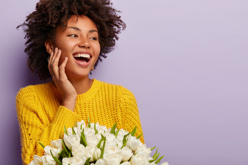 Wall Mural - Overjoyed pleasant looking dark skinned woman with curly hair, gazes aside, keeps hand on cheek, smiles broadly, wears yellow jumper, carries white spring flowers, isolated over purple background.