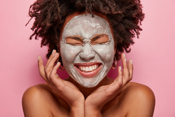 Wall Mural - Face care procedure concept. Gentle beautiful woman touches face, smiles broadly, shows white teeth, has clay mask on face, poses half nude, isolated over pink background. Anti aging concept