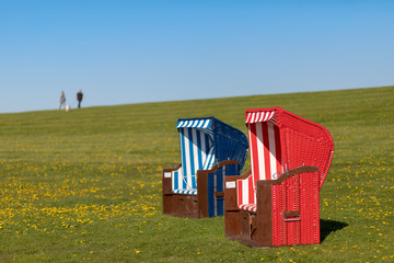 A blue and a red beach chair standing on the green meadow of the dike at the North Sea  with two persons blurred in the background