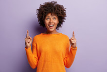 Positive Young Good Looking Woman With Afro Hairstyle, Indicates With Both Index Fingers, Wears Casual Orange Sweater, Feels Pleased, Isolated Over Purple Background. People And Promotion Concept