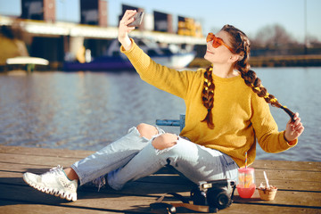 Wall Mural - Beautiful girl sitting on the pier near the river. Hipster model yellow with brunette long hair. The girl at hot sunny day taking pictures on phone camera, Summer vibes. Girl take a selfie. Drink