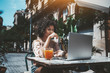 © skyNext - A curly-hair dazzling young African-American female freelancer is working with her project on the laptop remotely from a city cafe outdoors, with a glass of delicious fresh juice near on the table