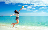 Fototapeta Morze - Happy young woman in casual style fashion and straw hat jumping at sand beach. Relaxing and enjoy holiday at tropical paradise beach with blue sky and clouds. Girl in summer vacation. Summer vibes.