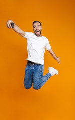 Wall Mural - Full length photo of optimistic guy 30s in casual wear laughing and taking selfie on cell phone, isolated over yellow background