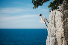 Beautiful Slim Dark Haired Young Woman In White Light Dress, With Long Legs Hangs On A Rope On The Rock Above The Sea, Looks Like Falling Or Dancing And Blue Sky And Sea Background.