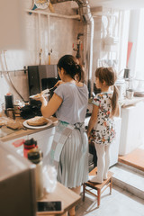 Wall Mural - Little girl staying on the stool next to her mother is cooking pancakes for the breakfast in the little cozy kitchen