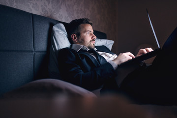 Businessman in suit lying in the bed in hotel room and using laptop for work. Overworking concept.