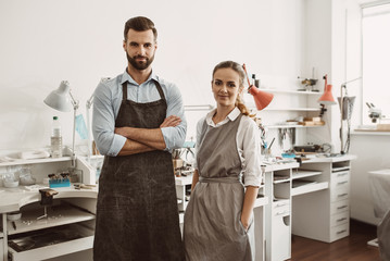 Business for a couple. Portrait of smiling couple in aprons standing at their jewelry workshop and looking at camera