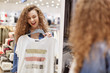 © sementsova321 - Curly girl with wide smile chooses t shirt in front of large store mirror. These clothes white with stripes in middle of three colors, golden red and silver. You can wear at beach and your home.