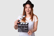 Movie concept. Positive young woman in hedgear holds cinema clapper, dressed in casual t shirt, has pleased expression, isolated over white background. Smiling actress prepares for shooting film