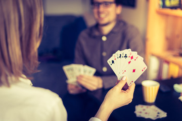 Friends are playing cards together at home. Woman is holding cards in her hands, man in the blurry background.