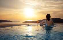 Woman In Infinity Swimming Pool At Golden Sunset. Girl Admiring Sunset From The Pool With A Cocktail Near. Summer Vacation Holiday