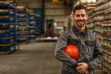Fototapeta  - Shot of a handsome young bearded factory worker in uniform holding protective hardhat smiling joyfully to the camera posing at the warehouse of a metalworking company