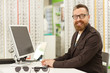 © Zoriana - Horizontal portrait of a cheerful mature male professional optometrist working at his eyewear store. Handsome male optician smiling to the camera, using his computer
