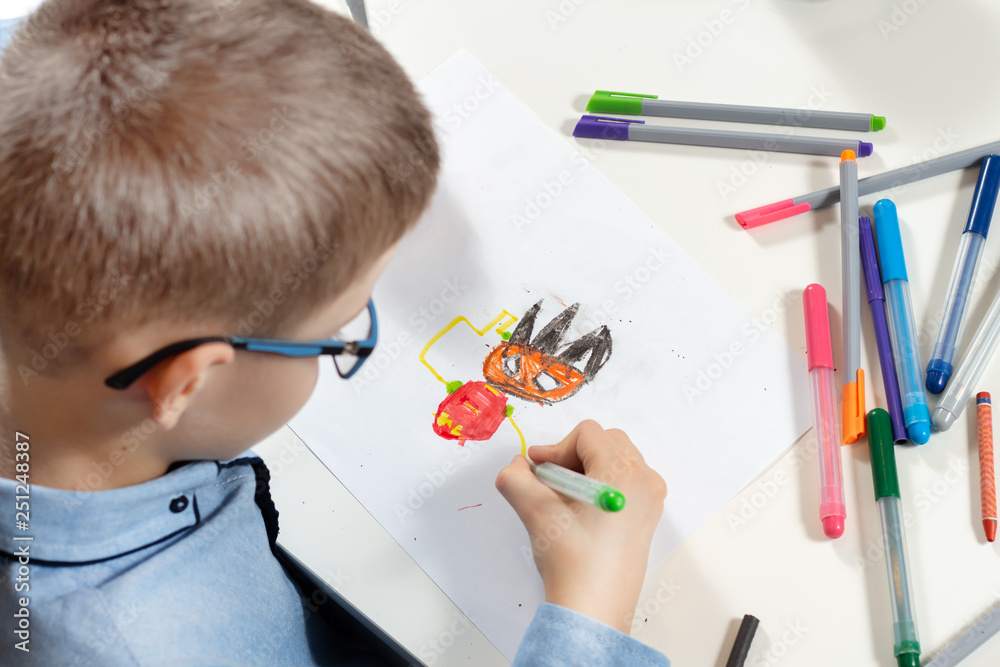 Chłopiec siedzi przy biurku i rysuje wymyśloną postać na białej kartce papieru. Kolorowe flamastry leżą dookoła na białym blacie. - obrazy, fototapety, plakaty 