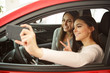 © Nestor - Two pretty girls making selfie in automobile cabin. Girls sitting inside, smiling, one girl holding phone. Female customers happy because of purchasing new auto in car center.