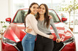 © Nestor - Font view of two girls buying new red auto in car dealership. Pretty female customers hugging  each other, leaning on new red car and smiling, posing at camera. Transportation concept.