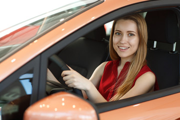 Smiling beautiful redhaired lady sitting in car on seat of driver, keeping hands on wheel, looking ar camera and posing. Happy customer testing new comfortable machine in car dealership.