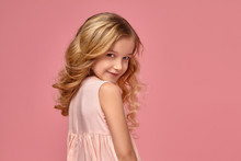 Little Girl With A Blond Curly Hair, In A Pink Dress Is Posing For The Camera