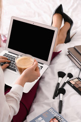 Young businesswoman with laptop drinking coffee on bed in morning