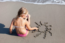 Kid Is Drawing Sun In Sand At Beach. Concept Of Children Picture, Sand Painting At Summer Vacation, Holiday And Travel. Little Girl Is Sitting In Waves Outdoors. Child Is Enjoying Summertime.