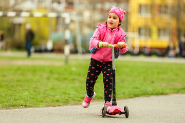 Wall Mural - Little child learning to ride a scooter in a city park on sunny summer day. Cute preschooler girl riding a roller. Kids play outdoors. Active leisure and outdoor sport for children.