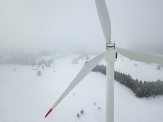 Aerial view of wind turbine in snow covered landscape in Swizterland. Tall pylon in fog, fir trees in the background.