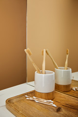 board with bamboo toothbrushes in holder and ear sticks beside mirror on white table and beige background