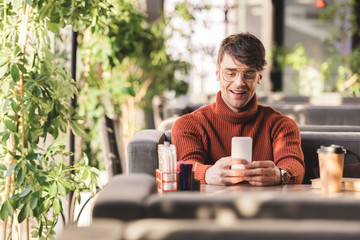 Sticker - selective focus of smiling man using smartphone near disposable cup and book in cafe