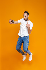 Wall Mural - Full length photo of young guy 30s in casual wear laughing and taking selfie on cell phone, isolated over yellow background