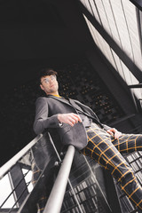 Wall Mural - young stylish man in glasses posing on stairs and looking at camera