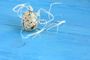 Quail Easter egg with shine decoration elements on spotted eggshell and natural straw nest with selective focus on blue wooden textured background with rough surface. Easter holiday card